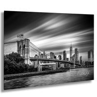 A black and white photograph of the Brooklyn Bridge and the New York City skyline, with the bridge spanning across the frame and the city's skyscrapers visible in the background.