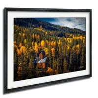 A framed photograph depicting a picturesque mountain landscape with a small wooden cabin nestled among the trees, surrounded by a vibrant display of autumn foliage.