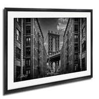 A black and white photograph of the Manhattan Bridge in New York City, with the bridge's intricate steel truss structure spanning across the frame between two tall buildings.