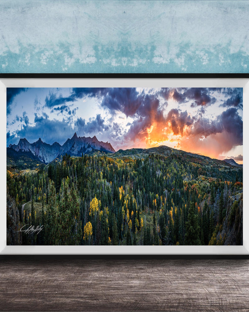 The image is a framed landscape photograph depicting a mountainous, forested area with a dramatic sky at sunset.