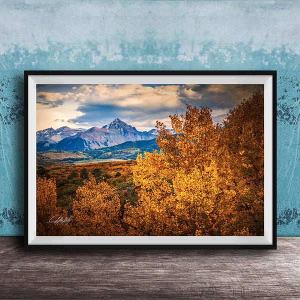 The image is a framed photograph depicting a scenic mountain landscape with a mix of autumn-colored trees and snow-capped peaks in the background.
