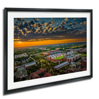 The image is an aerial view of a large sports stadium surrounded by a cityscape, with a dramatic sky in the background featuring clouds and a colorful sunset.