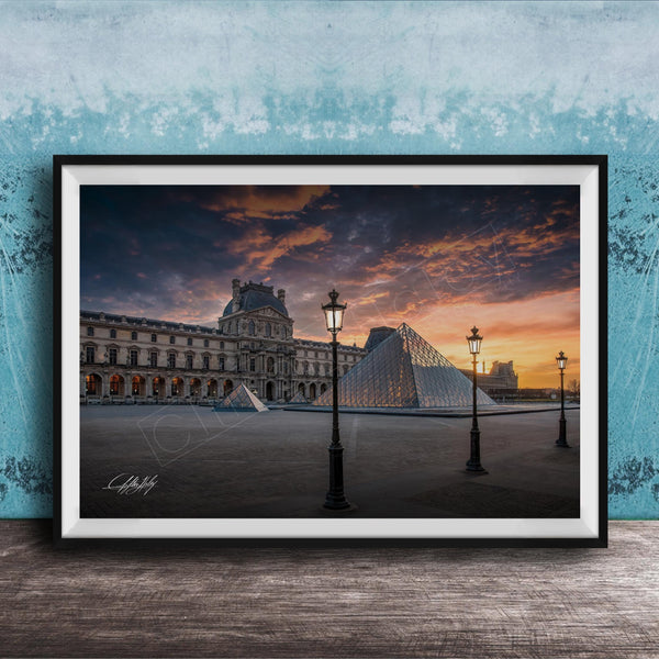 A framed photograph of the Louvre Museum in Paris, France, with the iconic glass pyramid structure of the Louvre Museum prominently featured against a dramatic sky at sunset.