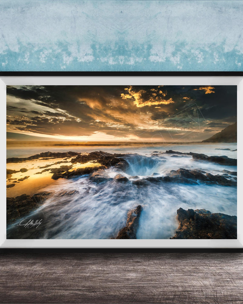 The image is a framed photograph of a rocky coastline with crashing waves, set against a dramatic sky with clouds and a sunset.