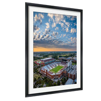 The image is an aerial view of a large sports stadium with a field, surrounded by buildings and a cloudy sky.