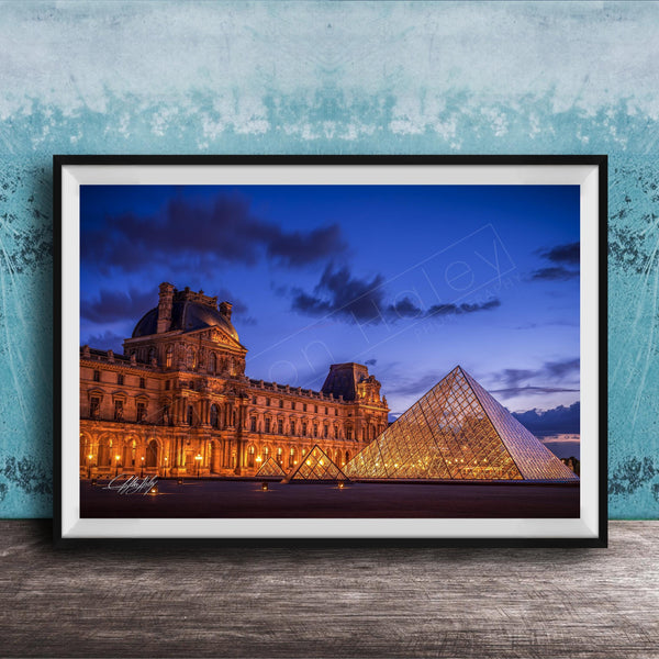 the Louvre Museum in Paris, France, with its iconic glass pyramid structure illuminated at night against a dark blue sky.