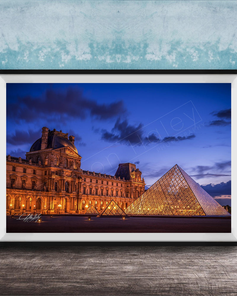 the Louvre Museum in Paris, France, with its iconic glass pyramid structure illuminated at night against a dark blue sky.