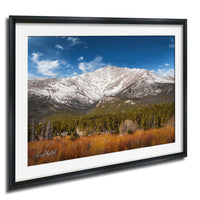 A framed photograph depicting a snow-capped mountain range with a forested valley below, set against a clear blue sky with wispy clouds.