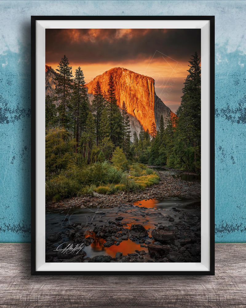 a breathtaking view of a mountain range at sunset, with the sun's warm glow illuminating the peaks and casting a vibrant orange and red hue across the sky. The foreground features a river flowing through the landscape, with rocks and trees lining its banks. The overall composition is visually striking and evokes a sense of tranquility and natural beauty.