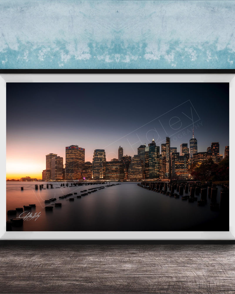 A framed photograph of the skyline of New York City at sunset, with the city's skyscrapers and landmarks illuminated against a dark blue sky.