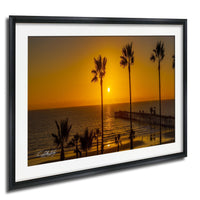 A framed photograph depicting a serene beach scene at sunset, with palm trees silhouetted against the warm, golden sky.