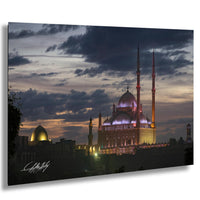 A large, illuminated mosque with multiple domes and minarets stands prominently against a dark, cloudy sky at dusk.