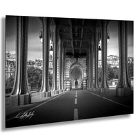 A black and white photograph of a long, arched bridge with columns and a central archway, leading to a cityscape in the background.