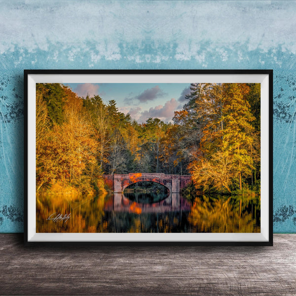 A framed photograph depicting a scenic autumn landscape with a red brick bridge spanning a tranquil lake, surrounded by vibrant fall foliage.