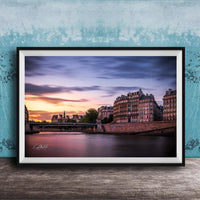 a picturesque cityscape at sunset, with a river running through the center and a row of buildings lining the riverbank. The sky is filled with warm, colorful hues, creating a serene and tranquil atmosphere. The buildings, with their unique architectural styles, add to the charm of the scene. The image is framed and displayed on a wooden surface, enhancing the overall aesthetic of the setting.