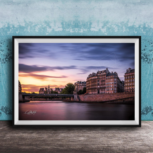 a picturesque cityscape at sunset, with a river running through the center and a row of buildings lining the riverbank. The sky is filled with warm, colorful hues, creating a serene and tranquil atmosphere. The buildings, with their unique architectural styles, add to the charm of the scene. The image is framed and displayed on a wooden surface, enhancing the overall aesthetic of the setting.