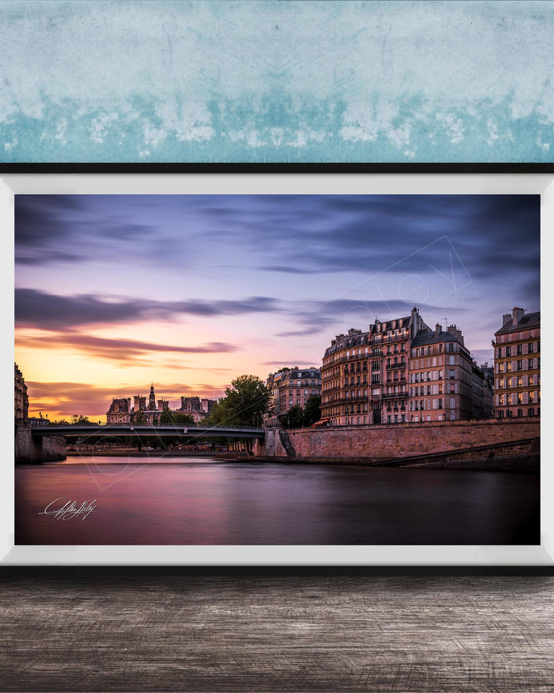 a picturesque cityscape at sunset, with a river running through the center and a row of buildings lining the riverbank. The sky is filled with warm, colorful hues, creating a serene and tranquil atmosphere. The buildings, with their unique architectural styles, add to the charm of the scene. The image is framed and displayed on a wooden surface, enhancing the overall aesthetic of the setting.