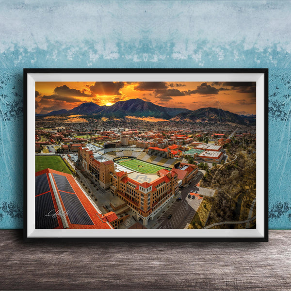 The image is a framed aerial view of a city with a large stadium, mountains in the background, and a dramatic sunset sky.