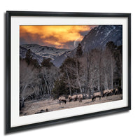 A framed photograph depicting a group of deer grazing in a snowy mountain landscape with a full moon and a vibrant sunset sky.