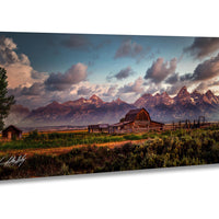 a serene rural landscape with a wooden barn and a small shed nestled in a grassy field, surrounded by trees and mountains in the distance. The sky is filled with clouds, creating a dramatic backdrop for the scene.