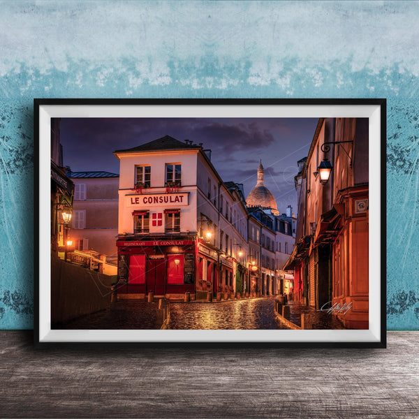 a charming street scene in Paris, France, with a row of colorful buildings lining the cobblestone street. The buildings are illuminated by warm street lamps, creating a cozy and inviting atmosphere. The sky above is a deep blue, providing a beautiful backdrop to the scene. The image is framed and displayed on a wooden surface, adding a rustic touch to the overall composition.