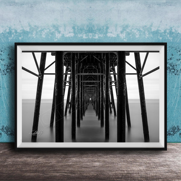 A black and white photograph of a long pier with multiple supporting columns, creating a sense of depth and perspective.