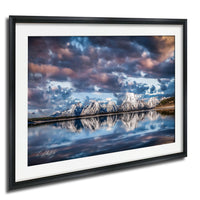 A framed photograph depicting a serene landscape with snow-capped mountains reflected in a calm lake, set against a dramatic sky with clouds.