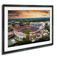 An aerial view of a large sports stadium with a crowd of spectators, surrounded by a cityscape and a dramatic sky with clouds and colors.
