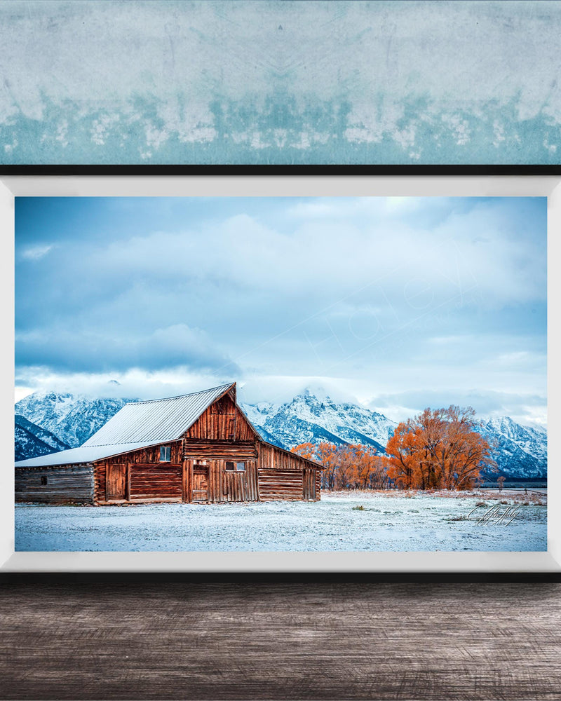 A large wooden barn with a sloping roof stands in a snowy field, with snow-capped mountains visible in the background.