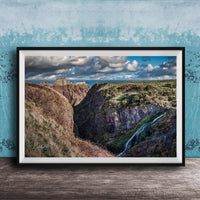 The image is a framed photograph depicting a scenic landscape with a waterfall cascading down a steep cliff face. The photograph is set against a blue wall, creating a striking contrast with the natural elements. The image showcases the beauty of the outdoors and the power of nature.