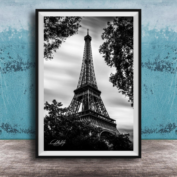 A black and white photograph of the Eiffel Tower, a famous landmark in Paris, France, framed and displayed on a wooden surface against a blue textured background.