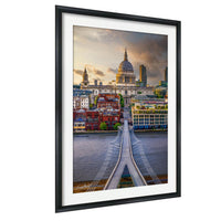 a cityscape with a large domed building in the background, surrounded by smaller buildings and structures. The scene is captured from a bridge over a river, with the bridge's railing visible in the foreground. The sky above is cloudy, creating a dramatic atmosphere. The image is framed, suggesting it is a piece of artwork or a photograph.