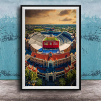 The image is an aerial view of a large stadium with a red roof, surrounded by buildings and a blue sky with clouds.
