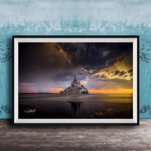 A framed photograph depicting the iconic Mont Saint-Michel island at sunset, with the island's reflection visible in the calm waters below.