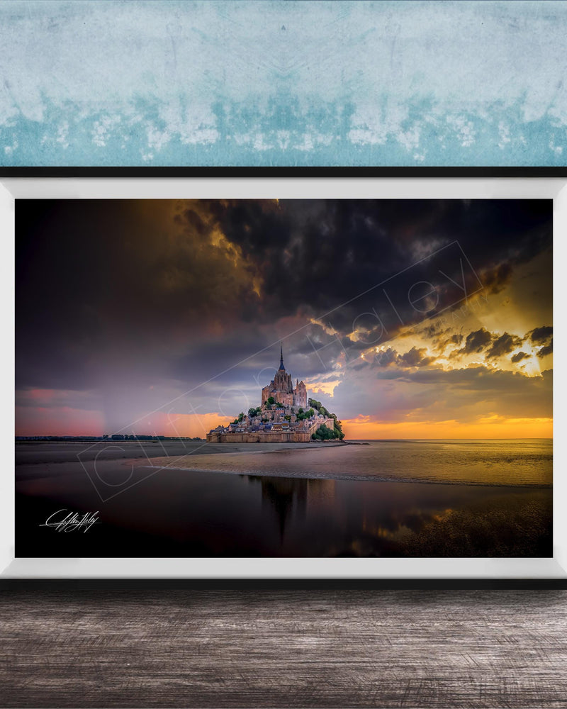 A framed photograph depicting the iconic Mont Saint-Michel island at sunset, with the island's reflection visible in the calm waters below.