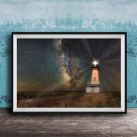 A framed photograph depicting a lighthouse standing in a field at night, with a starry sky and a galaxy in the background.