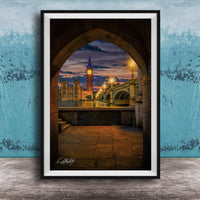 a cityscape at night, with a clock tower and a bridge visible in the background. The clock tower is illuminated, and the bridge is lit up, creating a warm and inviting atmosphere. The scene is framed in a black frame, which contrasts with the blue wall in the background. The image is a collage of multiple images, and the artist's signature is visible in the bottom right corner.