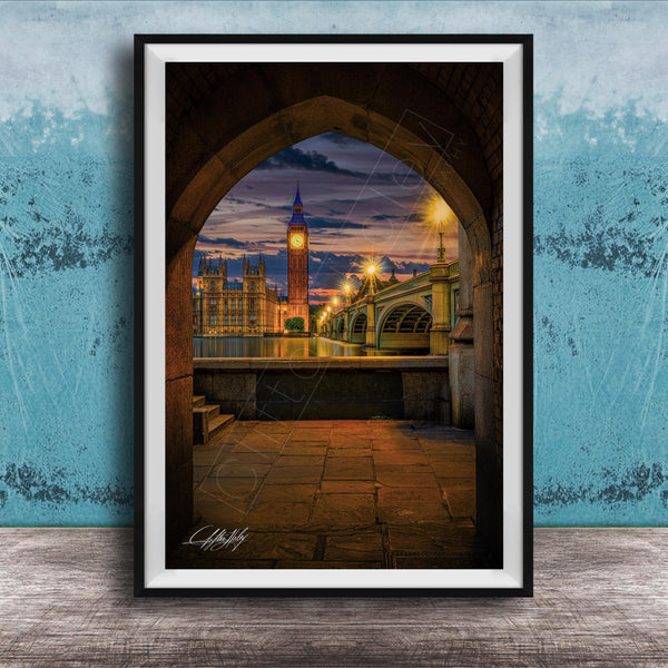 a cityscape at night, with a clock tower and a bridge visible in the background. The clock tower is illuminated, and the bridge is lit up, creating a warm and inviting atmosphere. The scene is framed in a black frame, which contrasts with the blue wall in the background. The image is a collage of multiple images, and the artist's signature is visible in the bottom right corner.