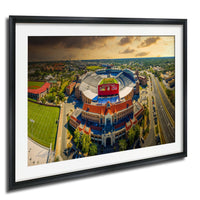 An aerial view of a large sports stadium with a tennis court and surrounding buildings, set against a cloudy sky.