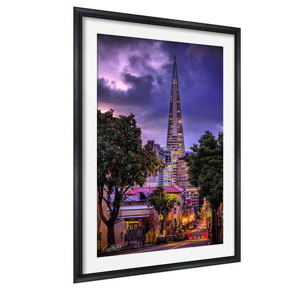 a cityscape at night, with a tall, illuminated skyscraper standing prominently in the background. The city is surrounded by trees and buildings, creating a vibrant and lively atmosphere. The sky above is filled with clouds, adding a dramatic touch to the scene. The image is framed, suggesting it is a piece of art or photography.
