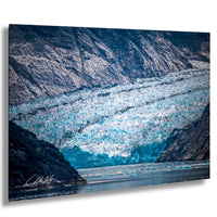 A large, majestic glacier with a blue-green hue dominates the scene, partially submerging the surrounding landscape. The glacier extends across the entire image, creating a striking contrast against the rocky terrain. The water below the glacier appears calm and serene, reflecting the natural beauty of the scene. The image is a captivating representation of the power and grandeur of nature.