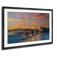 A framed photograph of the San Francisco Bay Bridge at sunset, with the bridge illuminated by warm, golden lights against a vibrant sky.