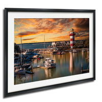 a serene harbor scene at sunset, with boats docked in the calm waters and a lighthouse standing tall in the background.