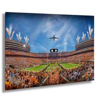 A large crowd of people in orange clothing gathered in a stadium, with a plane flying overhead and fireworks lighting up the sky.