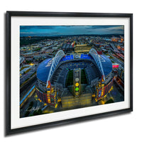 The image is a framed aerial view of a large stadium with a distinctive curved roof, surrounded by a cityscape at night.