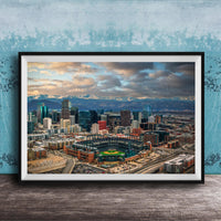 The image is a framed aerial view of a city skyline, featuring a baseball stadium and mountains in the background.