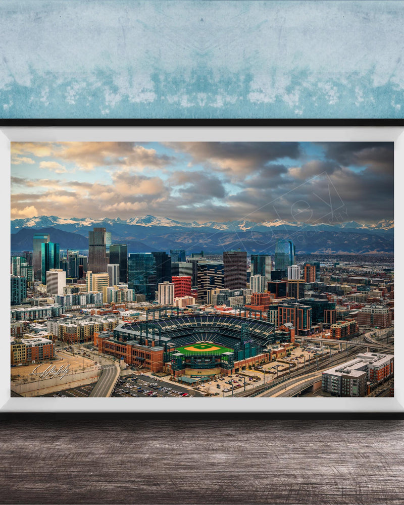 The image is a framed aerial view of a city skyline, featuring a baseball stadium and mountains in the background.