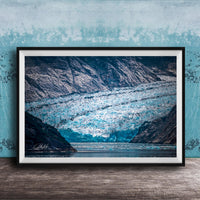 A framed photograph of a large, majestic glacier with a blue-green hue, set against a backdrop of rugged mountains and a serene body of water.