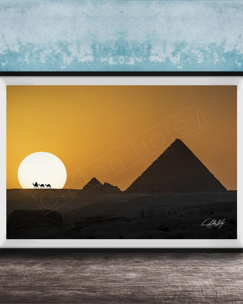 A framed photograph depicting the silhouette of the Great Pyramids of Giza against a vibrant sunset sky, with camels in the foreground.