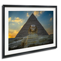 a large stone statue of a man with a headdress, standing in front of the iconic Great Pyramid of Giza in Egypt. The sky above the pyramid is filled with clouds, creating a dramatic backdrop for the scene. The photograph is framed and appears to be a high-quality image of the Great Pyramid and the Sphinx.